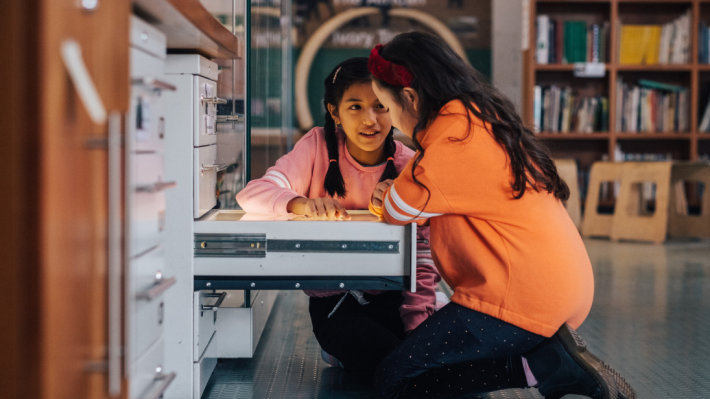 two girls, one in pink jumper and one in orange jumper, kneeling down looking into a museum collections drawer