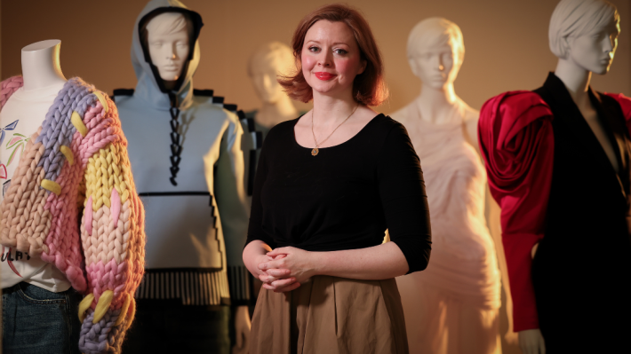 An art curator dressed in a black top and tan skirt stands in the centre of five white mannequins modelling fashion items from the Ulster Museum collection. Front left is a chunky wool cardigan, front right is a black blazer with large pink shoulder detail like a rose. Slightly in the distance to the left of the curator is a blue and black hoodie outfit and in the distance to the right is an all white draped dress. The face of the fifth mannequin peers over the curator's shoulder.