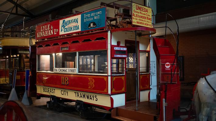 A red tram sits in a museum gallery. The upper deck is decorated with handdrawn advertisements.