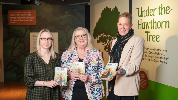 Three women standing side-by-side in front of cream and green exhibition graphics saying 'Under the Hawthorn Tree.' The woman on the left wears glasses and her hair is straight - she is holding a book by the same name, in Irish. The woman in the middle also wears glasses and shoulder length hair, also holding a copy of her book (she is the author). The woman on the right as short styled hair, she is wearing a neck scarf and cream blazer and she poses with an English cover of the same book.