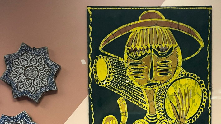 A view into a museum case. On the right, we can see the top half of a batik artwork, with a figure depicted in yellow wax on a black background.