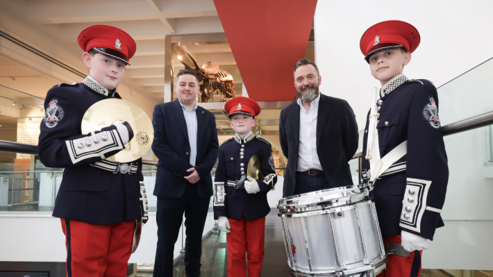 Three young band members dressed in their red and navy uniforms, with hats and white gloves, stand for a picture with two other people from the Belfast Bands Forum and National Museums NI. The boy on the left and boy in the middle are both holding cymbals, the boy on the right plays a side drum.