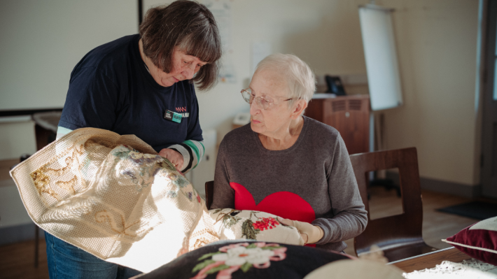 Two ladies, one with short dark hair and one with short white hair, studying stitching on some textiles. 