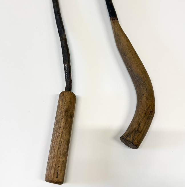 Two thatching forks lie on their sides. The handle of one is short and straight, while they other is long and curved.