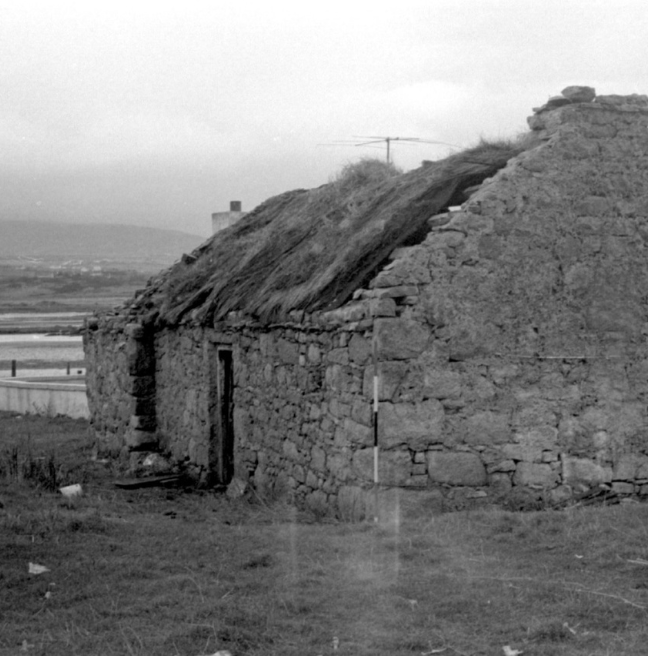 A black and white image of a derelict cottage with a thatched roof falling in. In the background we can see a body of water. 