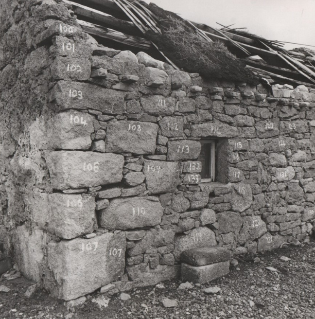 A black and white image of the walls of a run-down cottage. The stones are numbered with white chalk. 