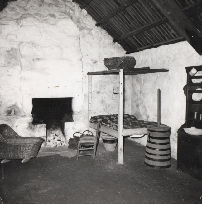 The inside of a byre dwelling staged with objects including a butter churn, a corner bed, a dresser with crockery, and a baby's cradle. 