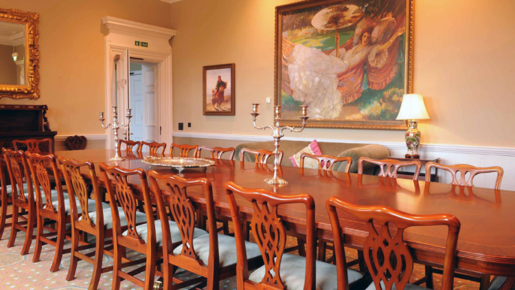 Long table and chairs in a dining room with two candelabras up the middle of the table. A large, colourful painting is on the wall behind and a lit lamp on the side table.