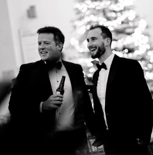 A black and white image of two men dressed in tuxedos, one is holding a bottle of beer, with a lit up Christmas tree in the background.