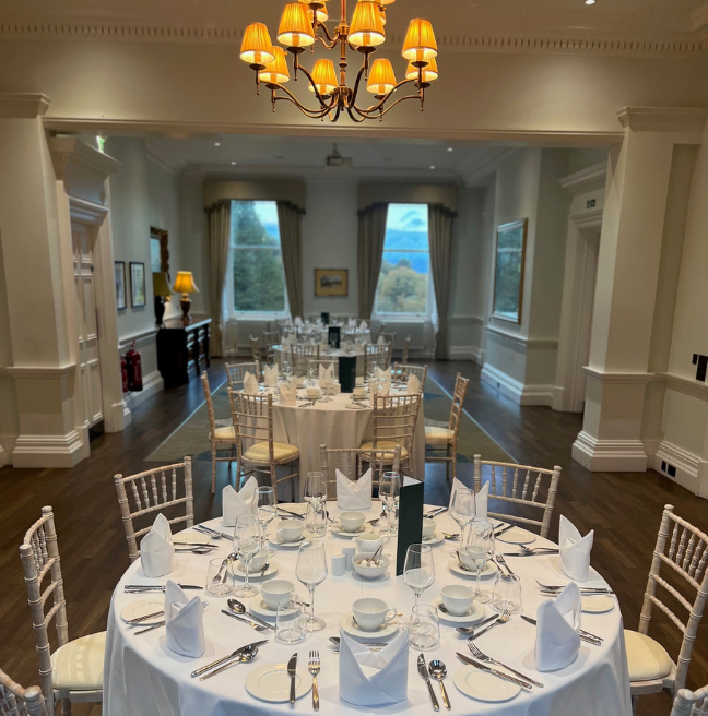 Three round tables laid out in the drawing room of Cultra Manor, all dressed in white cloth. Two large windows in the background let in natural light and a chandelier hangs above one of the table.