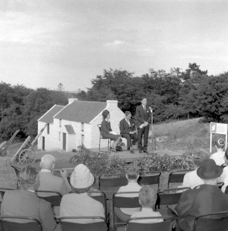 A black and white image of an event. In the foreground, a group of people are listening to a speaker on a podium. In the background, a farm sits on a sloping hill.
