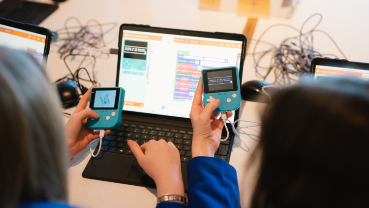 Two school pupils in front of a laptop holding game boy devices doing coding.