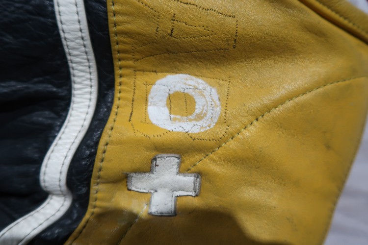 A close up of the top of a racing jumpsuit. There are white patches spelling 'O +' sewn on. The patches look like they are more closely adhereing to the jumpsuit than in previous photographs.