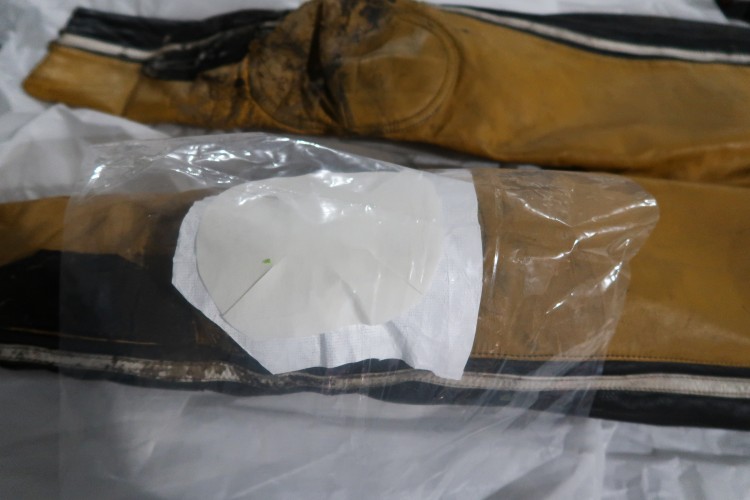 A close up of the knee of a yellow and black jumpsuit with a piece of white cloth over it. Over the cloth is a smaller piece of white cloth. Over that is a sheet of plastic.