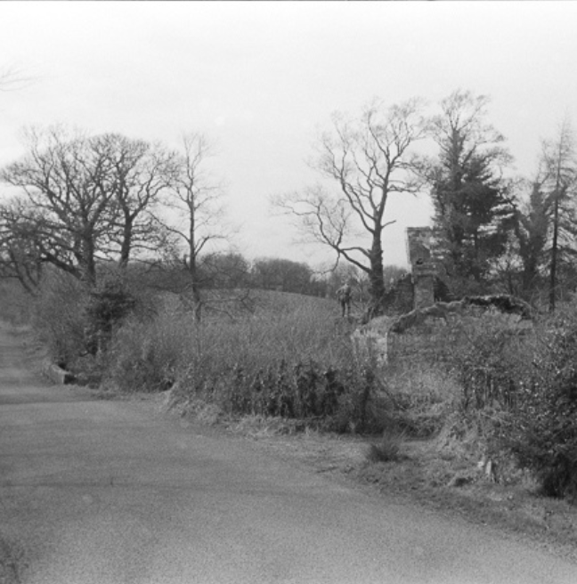 A black and white photograph of a dilapidated cottage by the side of a road.