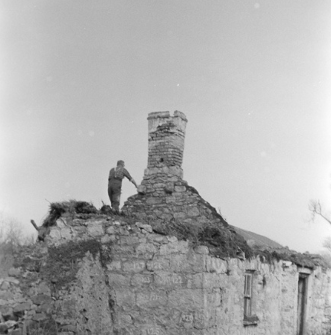 A black and white image of a man standing on a dilapidated cottage. He is holding on to the chimney, which still stands, albeit precariously. 