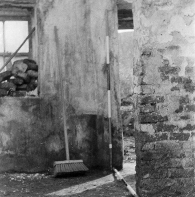 A black and white photograph of the interior of a rundown cottage. There is a large jamb wall jutting out from the right. 