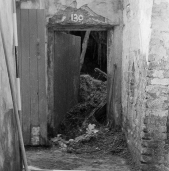 A black and white photograph of the entrance way of a rundown cottage. There is a large jamb wall on the left, a doorway to more interior rooms ahead, and to the left is the door to the cottage. 
