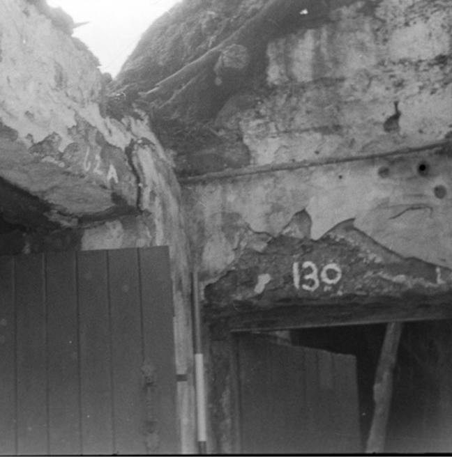 A black and white photograph of the interior of a rundown cottage. The focus is on the upper walls where two rooms ajoin. 
