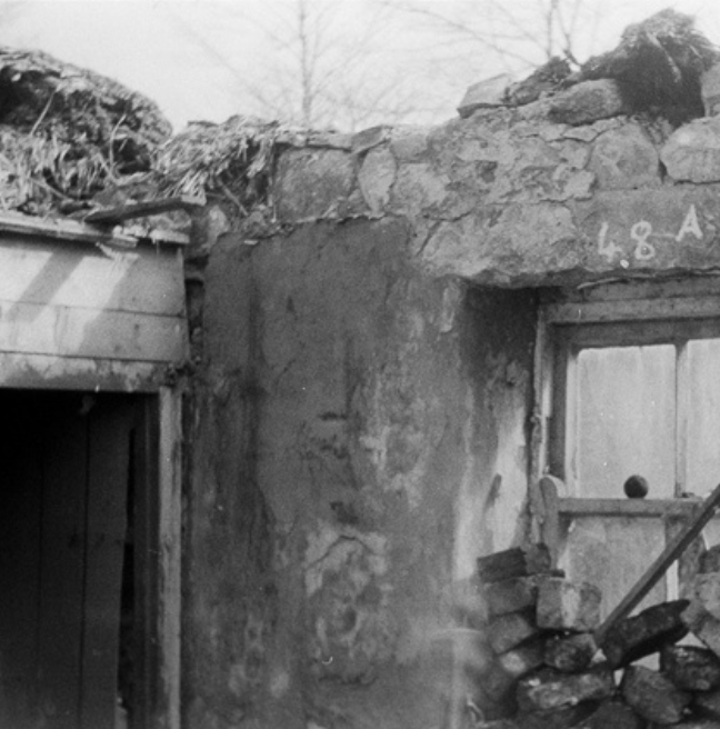 A black and white photograph of the interior or a rundown cottage showing the main room and a wall that has been raised to add more privacy. While the walls are brick, the privacy wall seems to be made of wood.