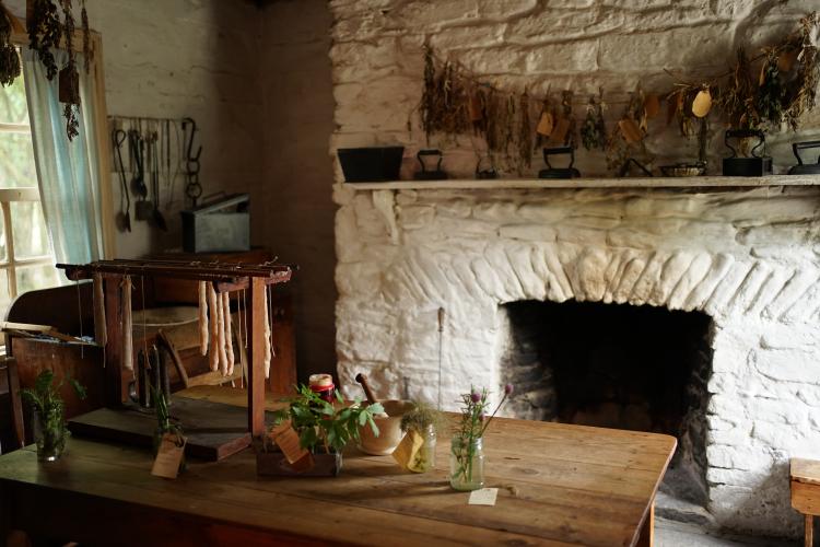 A room in a log farmhouse, with a large fireplace at the rear and a table in the centre, topped with herbs and candles setting.