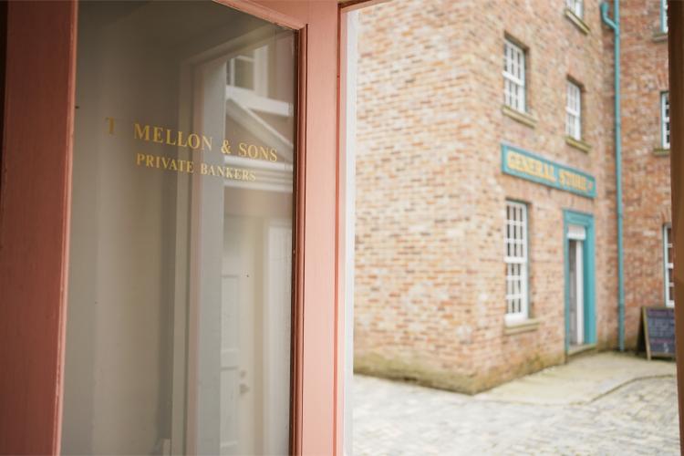 An image of the door of a bank, reading 'T. Mellon & Sons, Private Bankers'. Out the door, we can see a cobbled main street and a red brick building.