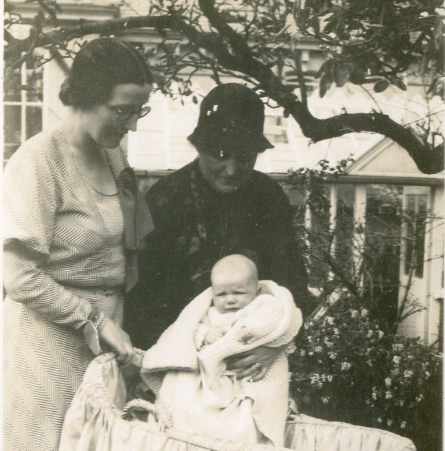 A black and white photograph of two woman, one older than the other, standing over a pram. The elder woman holds a baby in her arms, facing the camera.