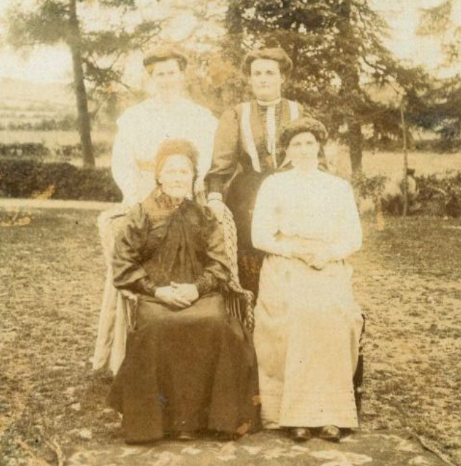 An old black and white photograph of four women posing for a photograph in a garden. Their dress suggests this photograph is from the early 1900s.