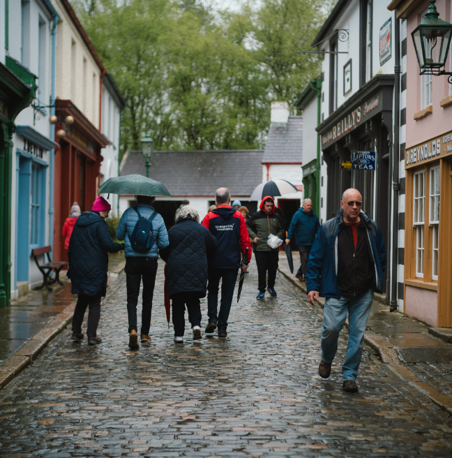 A street in Ulster American Folk Park with a crowd of people walking in the rain with an umbrella 