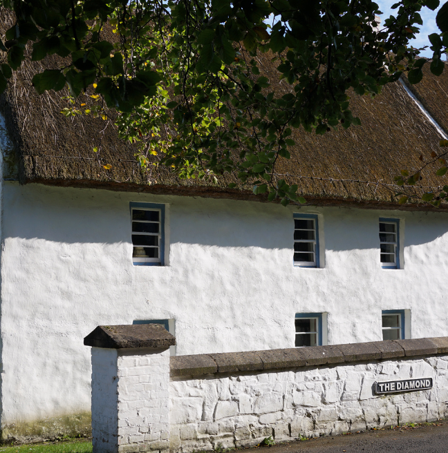 A whitewashed cottage with a thatch roof stands in the sun.
