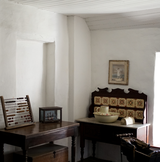 The interior of a cottage bedroom. A side table with a wash basin is visible. 