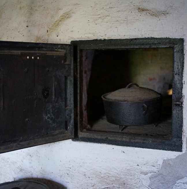 An oven set deeply into the wall is seen with its door open. An iron pot is inside.