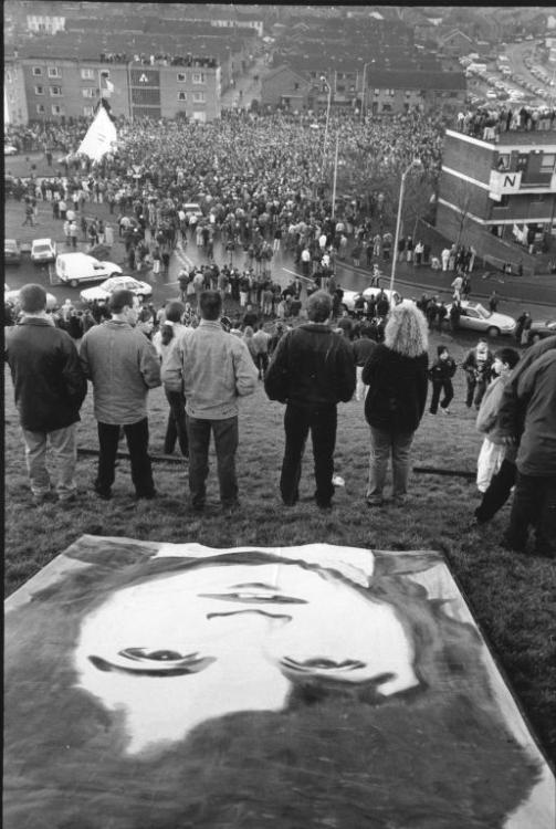 Black and white photograph of Bloody Sunday commemorations, large print of a person on a field, can see the back of crowds standing