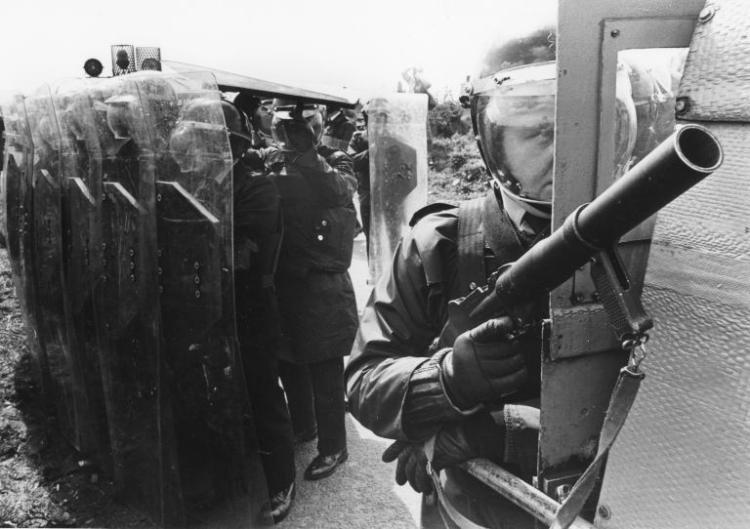 Black and white photograph of police man at rear doors of landrover with rubber bullet gun, with line of policemen behind with riot shields at funeral of hungerstriker Tom McIlwee