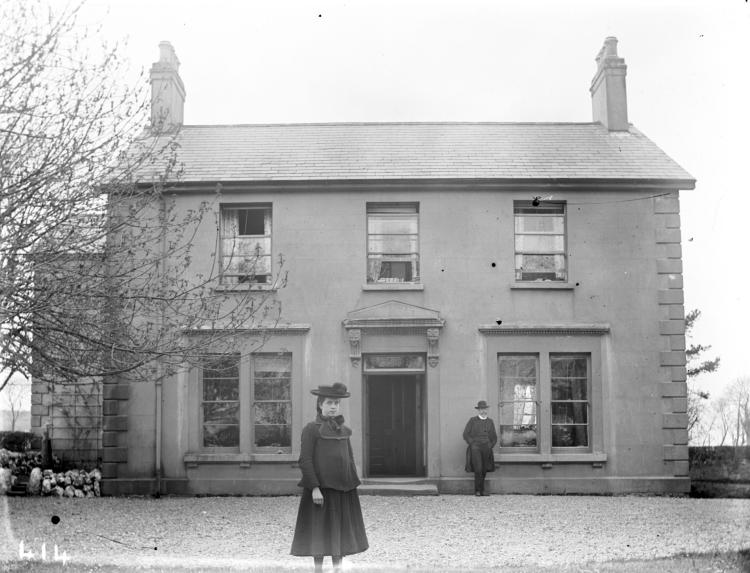 A woman stands outside a lovely two-storey home in a black and white photograph.