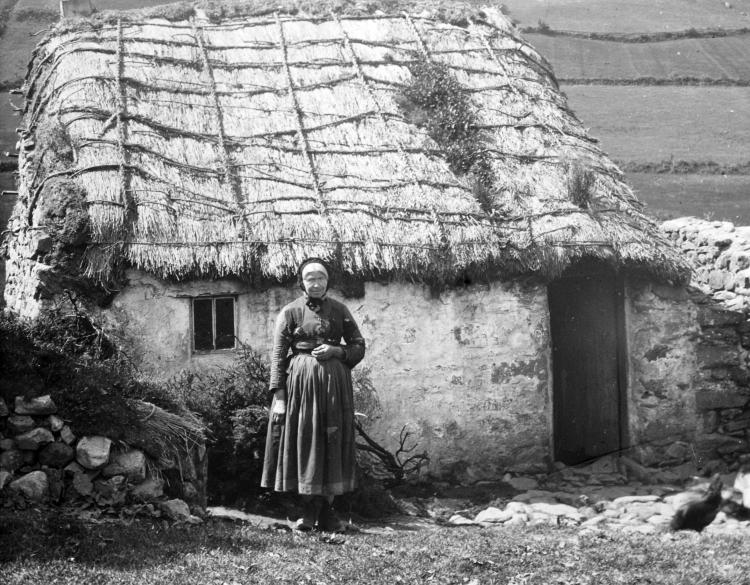 A woman stands outside a small thatched cottage in a black and white photograph.