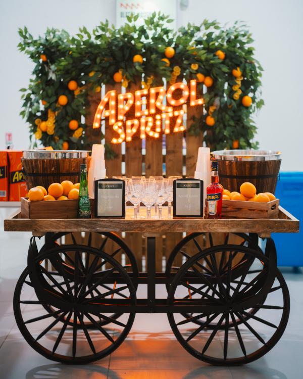 A cart with fruit and drink on it, with an orange lit up sign behind it saying Aperol Spritz. The sign has a foliage and oranges installation around it too.