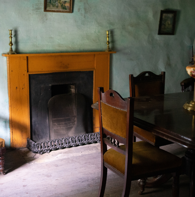The interior of a farmhouse showing a light wooden mantle around a fireplace.