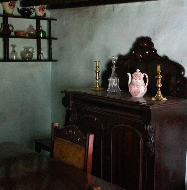The interior of a farmhouse showing a dark wooden cupboard with candlesticks and a jug on top.