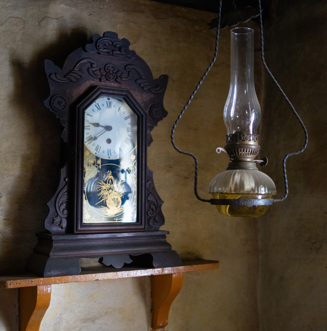 The corner of a farmhouse living room showing a clock and an oil lamp.