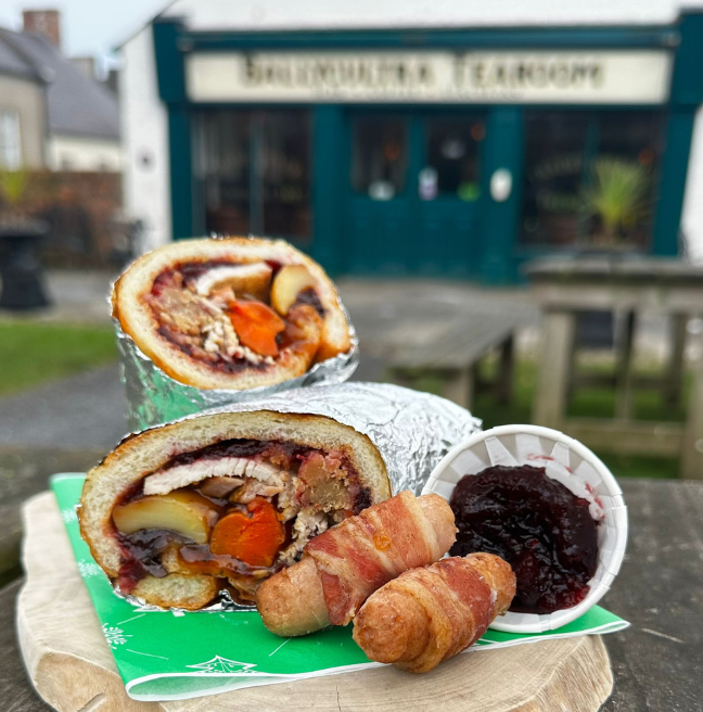 A picture of a Christmas food wrap, pigs in blankets and a pot of cranberry sauce all sitting on top of a wooden board with Ballycultra Tearoom, which is the Ulster Folk Museum cafe, blurred in the background.