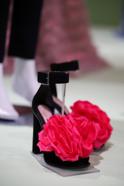A pair of black, block heel shoes, with an ankle strap and large pink rose detail on the toes, mounted in an exhibition display.