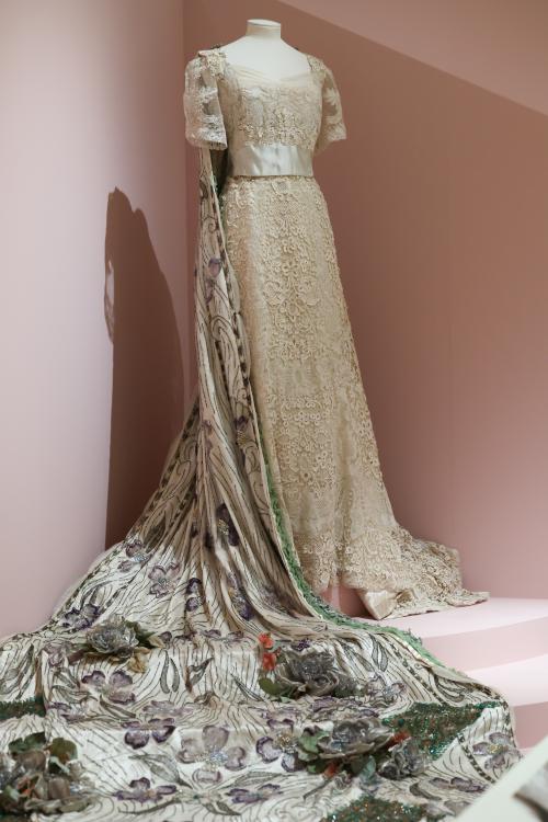 An ivory coloured dress with detailed train on a mounted in an exhibition display, set against a faded pink backdrop. The dress is made of Brussels lace and Irish crochet.
