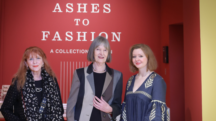 Three curators, all female, standing in front of a deep red coloured wall with white text on it saying Ashes on one line, to in the middle line, then fashion underneath that on another line. There are vertical white lines going down to the wall as well as part of the design.