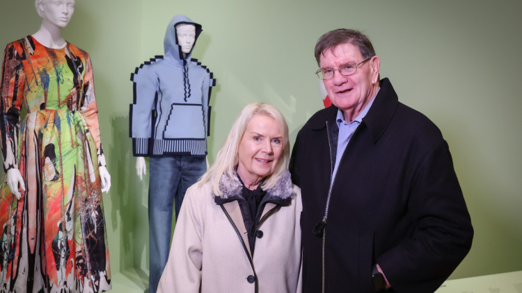 A couple standing in front of two outfits on mannequins, against a mint green wall.