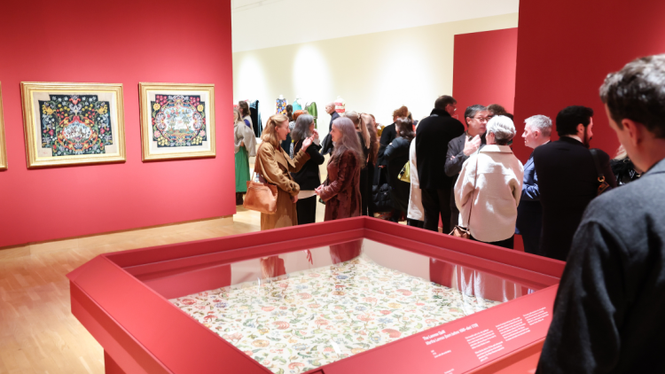 A wide shot of a gallery space, with deep red colour walls and people gathered in the background. The focal point of the photo is a wooden case with glass top containing a delicate, handstitched quilt from 1712.