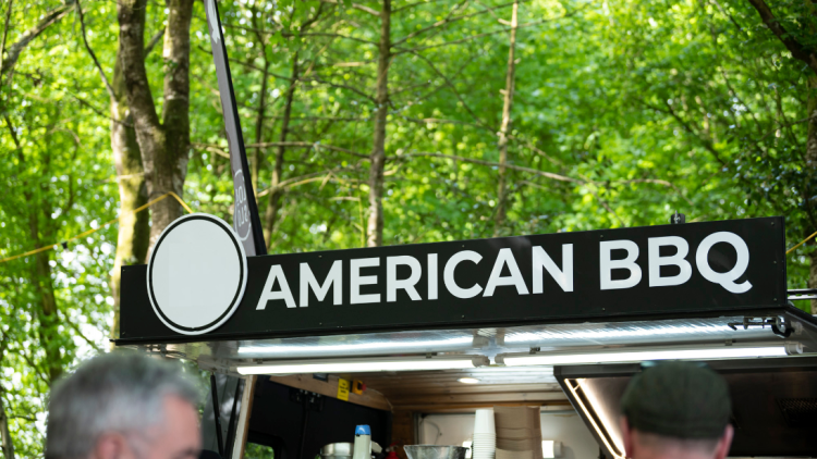 An outdoor food van saying American BBQ in white text on a black sign. Set against green trees in the background and you can just see the tops of some people's heads.