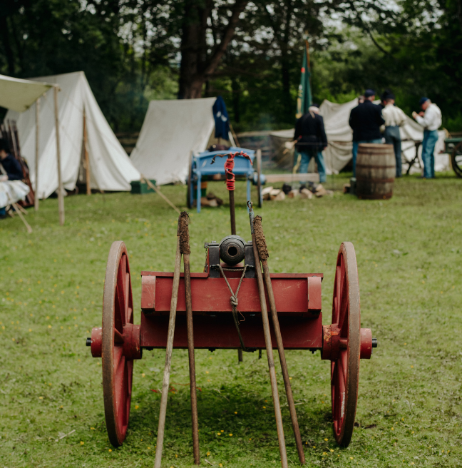 An old fire canon sitting in a field with white tents visible in the background. It's a staged setup of a scene from the American Civil War