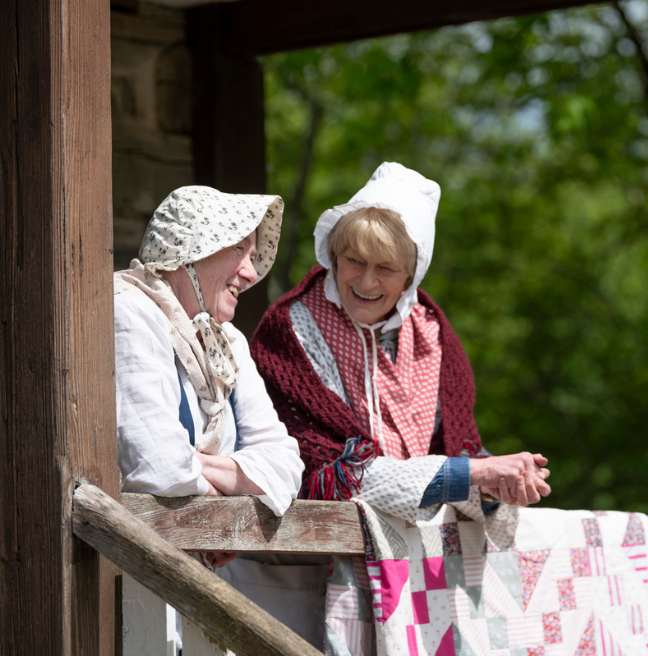 Two women dressed in American Frontier dress leaning on a wooden balcony which has a patchwork quilt draped over the edge too. They're smiling and appear to be laughing at each other and their conversation.