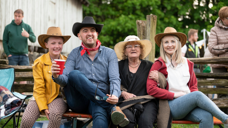 A family, all wearing cowboy hats and sat down on deck chairs at an outdoor music event, pose and smile for the camera.